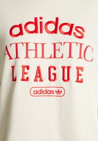 Witte katoenen T-shirt met opvallende rode tekst: "adidas ATHLETIC LEAGUE" en een logo. Het ontwerp is eenvoudig met een ronde hals.
