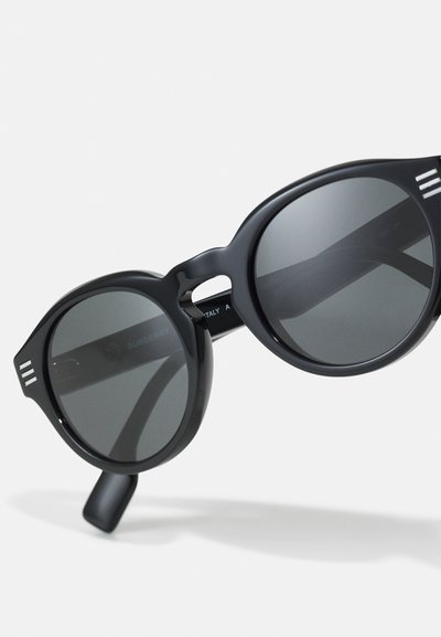 Occhiali da sole neri rotondi con lenti grigie, montatura in plastica spessa, con dettagli in argento sulle tempie e un design leggermente curvo.