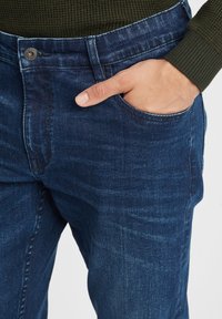 Main reposant dans la poche avant d'un jean en denim bleu foncé porté avec un pull en laine vert olive.