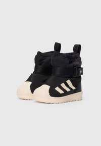 Bottes noires pour tout-petits avec des orteils en caoutchouc beige, des bandes Adidas beiges sur les côtés, des sangles noires ajustables et des tirants sur un fond blanc.