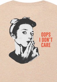 T-shirt color pesca chiaro con una grafica in bianco e nero di una persona con una fascia in testa, e il testo "OOPS I DON’T CARE" in rosso in grassetto.