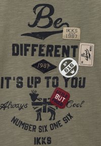 Zielona oliwkowa tkanina z czarnym napisem "Bądź Inny", łatami z napisem "IKKS od 1987", "Nr 616" oraz "Mały, ale Dumny", oraz stylizowanym psem.