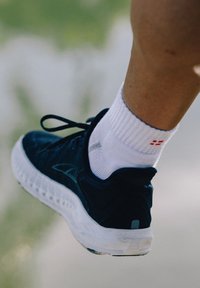 Czarny but sportowy z białą podeszwą, teksturowaną cholewką i czarnymi sznurowadłami. Noszony z białą skarpetką z czerwonymi akcentami.