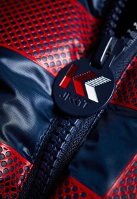 Takki, jossa on laivastonsininen pohja ja punaiset rei'itetyt yksityiskohdat, vetoketju mustalla logomerkillä sekä teksturoitu kangas, jossa on sileä pinta.