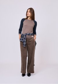 Pantaloni lunghi grigio-marrone con gambe larghe in tessuto di velluto a coste, abbinati a una maglietta a maniche lunghe beige e a una camicia a scacchi blu annodata in vita.