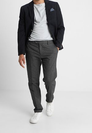 Uomo con blazer scuro, t-shirt bianca, pantaloni grigi su misura e sneakers bianche, che cammina su uno sfondo chiaro uniforme.