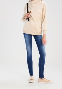 Sweat à capuche beige clair à coupe ajustée avec cordon de serrage, associé à un jean skinny bleu délavé et des chaussures beige à enfiler.