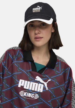 Bordeaux trøje med blå og grøn geometrisk mønster, med en sort krave, og en sort kasket med "PUMA KING" logo i hvid og guld.
