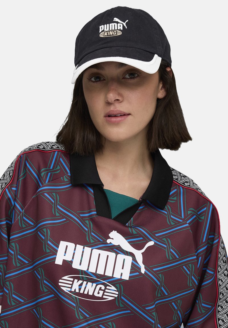Borvörös mez kék és zöld geometriai mintával, fekete gallérral, és egy fekete sapka "PUMA KING" logóval fehér és arany színben.