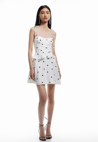 Biała sukienka peplum w czarne kropki, z cienkimi ramiączkami i dopasowanym gorsetem. Spódnica ma rozkloszowany krój i subtelną teksturę.
