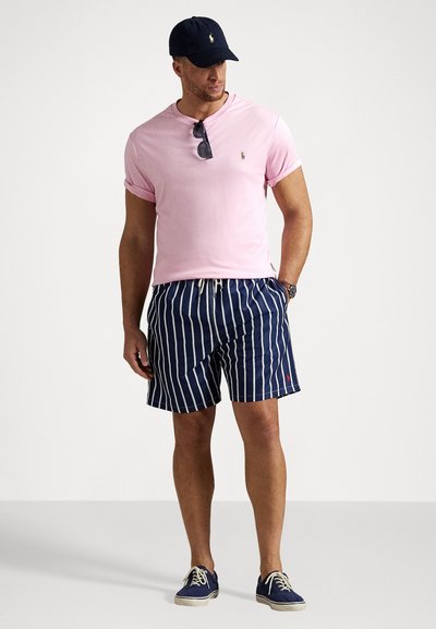 Rožinė trumpa rankovė t-shirt, tamsiai mėlynai dryžuoti maudymosi šortai, tamsiai mėlyna kepurė. Textūruotas audinys su maža logotipo etiketė ant marškinėlių ir šortų. Kasdieniniai batai.