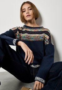 Granatowy sweter z dzianiny z kolorowymi geometrycznymi wzorami wokół dekoltu i mankietów, o luźnym kroju i długich rękawach.