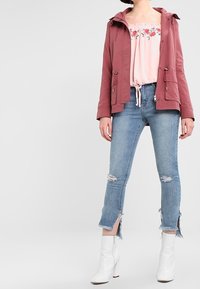 Donna che indossa un top off-shoulder floreale rosa, una giacca rosa polveroso, jeans cropped strappati e stivaletti bianchi con tacco.