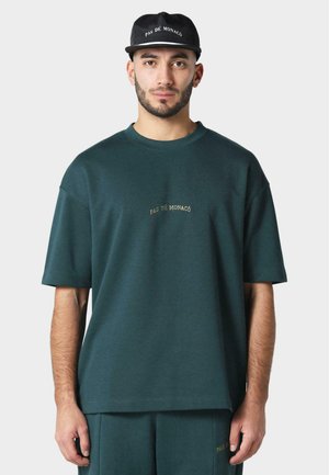 Dark green oversized t-shirt with short sleeves, featuring gold embroidery reading "PAS DE MONACO." Paired with matching sweatpants.