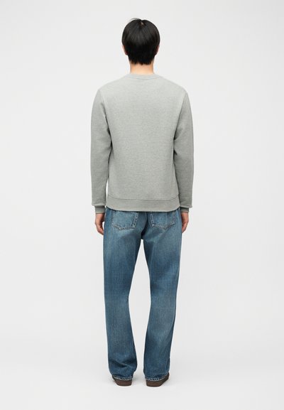 Άτομο που στέκεται γυρισμένο με την πλάτη φοράει απλό ανοιχτό γκρι φούτερ, φαρδιά μπλε τζιν και καφέ παπούτσια σε λευκό φόντο.