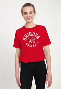 Young woman wearing a red T-shirt with "Shibuya 1961 Tokyo Baseball" text, paired with black pants, standing against a plain white background.