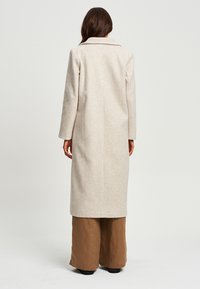 Lungo cappotto beige realizzato in morbido misto lana, con colletto a punta, taglio dritto e tasche laterali. Indossato sopra pantaloni larghi marroni.