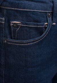 Poche de jean en denim bleu foncé avec des coutures en cuivre et trois rivets noirs sécurisant les bords du tissu.