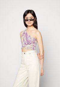 Top cropped assimétrico em padrão de espiral roxo, amarelo e teal. Feito de tecido elástico. Combinado com calças brancas de cintura alta.