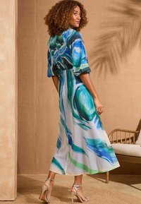 Maxi-jurk met abstracte blauwe en groene swirlpatronen, lichtgewicht stof, driekwart mouwen en een relaxed fit. Transparante hakken.