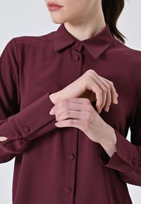 Burgundy long-sleeve shirt with a pointed collar, front button closure, and circular cuff buttons. Fabric appears smooth and lightweight.