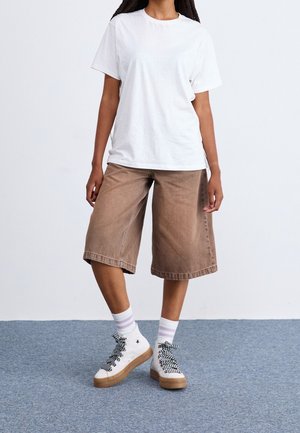 Person iført en helt hvid t-shirt, løse brune knælange shorts, hvide sokker med lyse striber og hvide høje sneakers med mønstrede snørebånd.