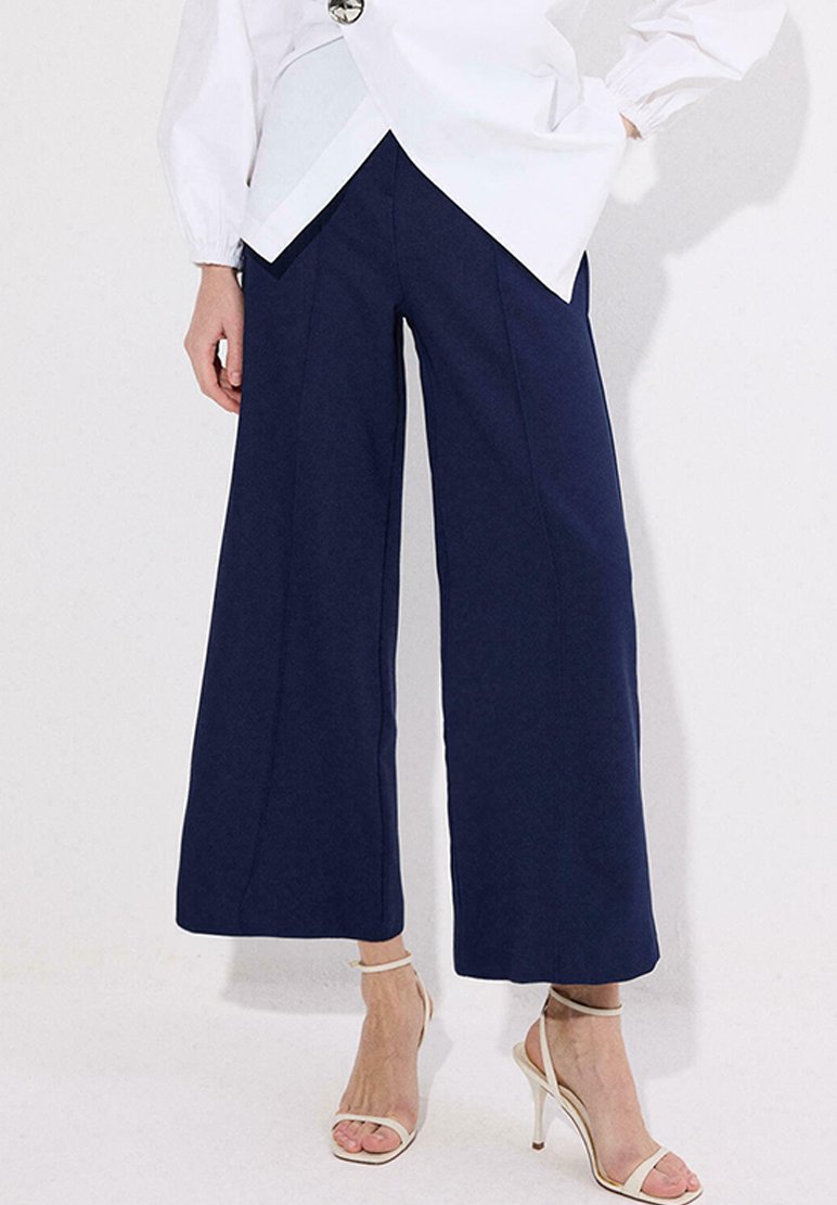 Γυναίκα που φοράει navy παντελόνι με φαρδιά μπατζάκια μέχρι τον αστράγαλο, λευκό πουκάμισο και μπεζ ψηλοτάκουνα σανδάλια, στέκεται μπροστά σε έναν απλό φωτεινό φόντο.