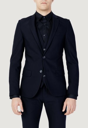 Completo blu scuro con giacca aderente dalle rever a scendere, pantaloni slim e chiusura a doppio bottone. Indossato sopra una camicia nera.
