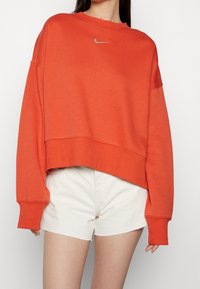 Pomarańczowa krótka bluza z bawełny z długimi rękawami, z małym srebrnym logo Nike na piersi, w połączeniu z białymi szortami.