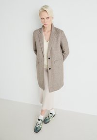 Hnedý kabát z vlnenej zmesi s klopenými chlopňami, dvomi prednými vreckami, skombinovaný s krémovým svetrom s v-výstrihom a svetložltou sukňou, doplnený zelenými teniskami.