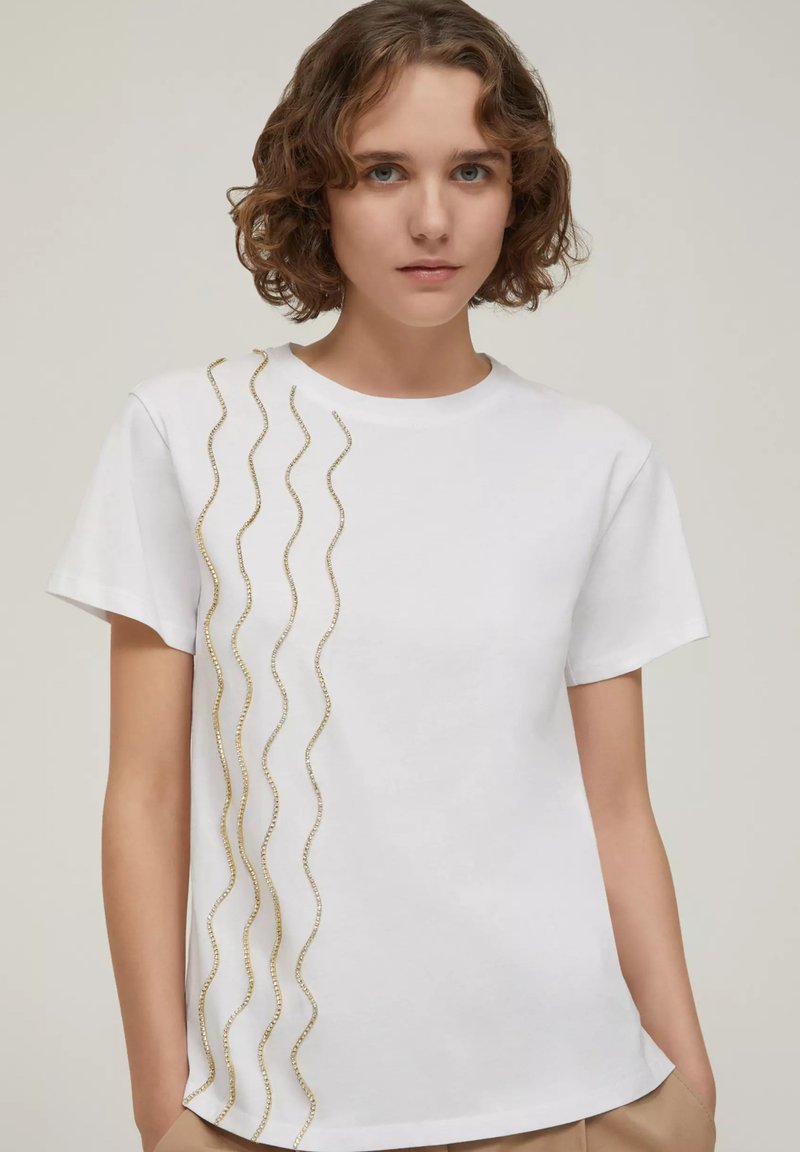 T-shirt bianco in cotone con dettagli in paillettes dorate ondulate su un lato. Collo rotondo, maniche corte e vestibilità leggermente rilassata.