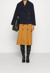 Manteau croisé navy raccourci, robe boutonnée couleur moutarde et bottes noires ajustées hauteur genou. Sac à main noir à la main.