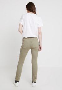 Pantalones cargo caqui con puños elásticos, dos bolsillos laterales y un corte relajado, combinados con una camiseta blanca sencilla en un entorno minimalista.