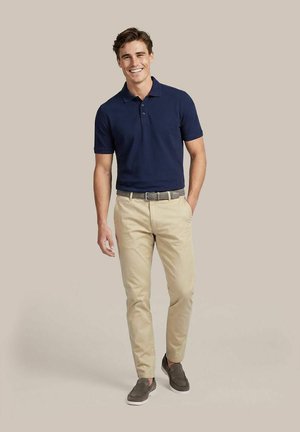 Man glimlacht, draagt een marineblauw poloshirt, beige broek met riem en grijze instapschoenen, staat tegen een beige achtergrond.