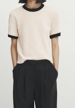 Woman wearing a short-sleeve beige textured top with black trim paired with high-waisted black wide-leg trousers, hands in pockets.