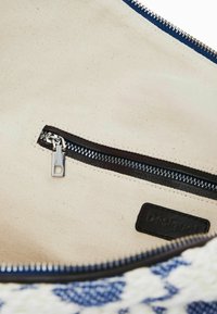 Sisätila kanvaskassissa, jossa on kermainen vuori, laivastonsininen vetoketju ja pieni mustasta nahasta valmistettu etiketti, johon on painettu "Desigual".