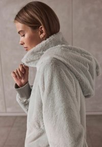 Lichtgrijze, donzige jas met capuchon en een zachte textuur. Heeft een hoge kraag en elastische manchetten, ontworpen voor comfort en warmte.