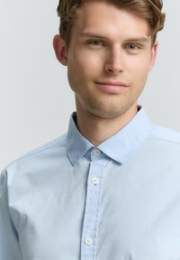Jaunas vyras su šviesia oda, trumpais šviesiai rudais plaukais ir mėlynomis akimis, dėvi šviesiai mėlyną marškinių mygtuką, šiek tiek šypsosi prieš neutralų foną.