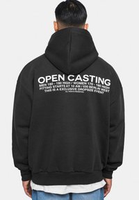 Schwarzer Kapuzenpullover mit großen weißen Schriftzügen auf dem Rücken, die "OPEN CASTING" und Einzelheiten zur Veranstaltung anzeigen. Weicher Stoff, lockere Passform.
