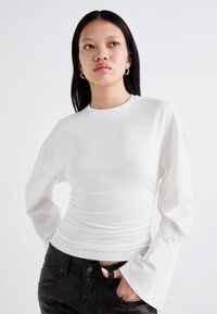 Long-sleeve white top with a fitted silhouette and ruched detailing, paired with black pants featuring a textured finish and standard pocket design.