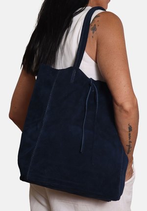 Frau mit langen dunklen Haaren, die ein weißes ärmelloses Oberteil trägt und eine große marineblaue Wildleder-Tasche über der Schulter trägt, sichtbare Tattoos am Arm.