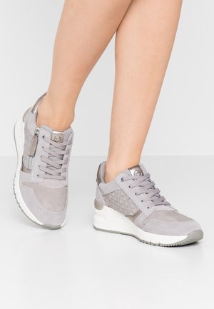 Legs wearing light gray lace-up sneakers with side zipper, geometric quilted pattern, and white thick sole on smooth gray floor.