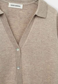 Cardigan beige lavorato a maglia con scollo a V, colletto a coste e chiusura a bottone. Presenta una consistenza morbida e un singolo bottone di colore chiaro.