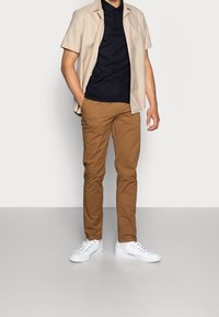 Beige kortärmad skjorta, marinblå polo, bruna byxor och vita sneakers som bärs av en person som står mot en neutral bakgrund.