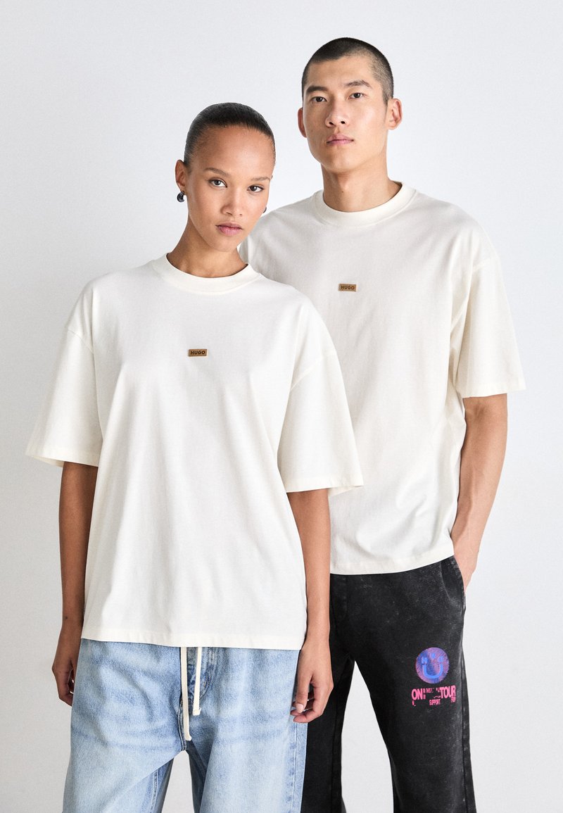 Kræmfarvede oversized bomulds t-shirts har et lille brunt logo mærke. Modellen til venstre bærer denimjeans, og modellen til højre bærer mørke bukser.