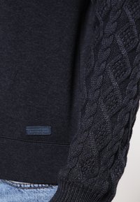 Mörk marinblå tröja med en texturerad flätad stickdesign på ärmarna och en slät kropp. Liten märkesetikett i botten.