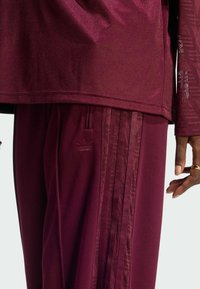 Survêtement athlétique bordeaux comprenant un haut texturé et un pantalon assorti avec trois bandes. Tissu lisse avec un léger éclat et détail de marque.