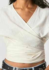 Biała bluzka w lekkim materiale, z dekoltem w serek i skręconym wzorem w talii. Połączona z dżinsami i dekoracyjnym paskiem.