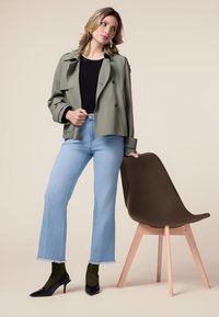 Chaqueta de doble botonadura verde oliva, top negro, vaqueros acampanados azules claros con dobladillo deshilachado, calcetines verdes, tacones negros, silla marrón con patas de madera.