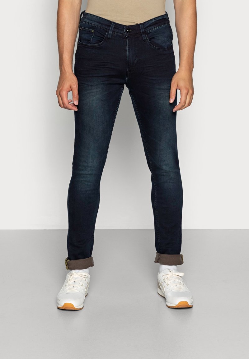 Muž v tmavě modrých slim fit džínech s ohrnutými lemy a bílých teniskách stojí na šedé podlaze proti bílé zdi.